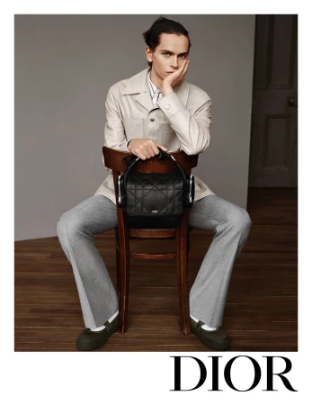 Model wearing a light beige jacket and grey trousers seated on a wooden chair holding a black quilted handbag against a neutral wall