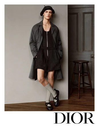 Fashion shot of a slender adult male model wearing a black romper suit with an overcoat, Mary Jane shoes and a black beret in a studio.
