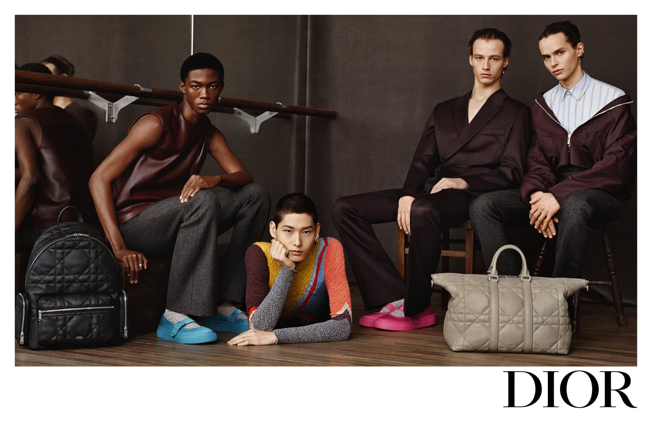 Fashion editorial shot of four male models wearing contemporary clothing, posing indoors with diffused lighting in a studio setting. 