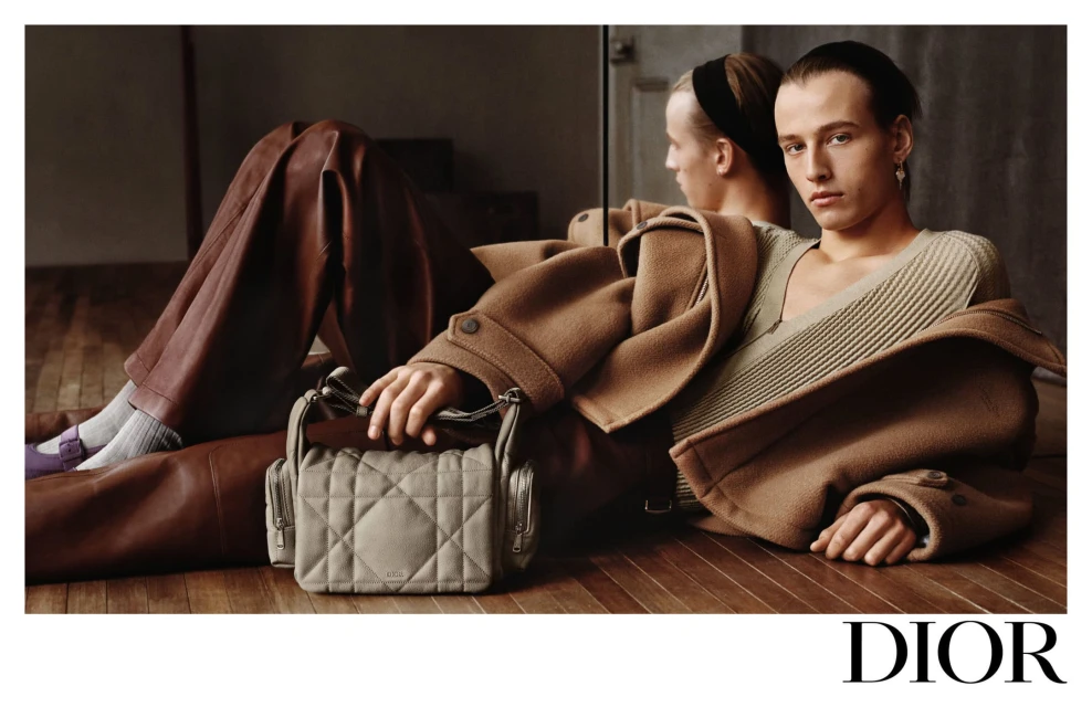 Fashion shot of a young adult male model in a tan coat with a neutral ribbed shirt, brown pants, and a gray handbag by Dior.