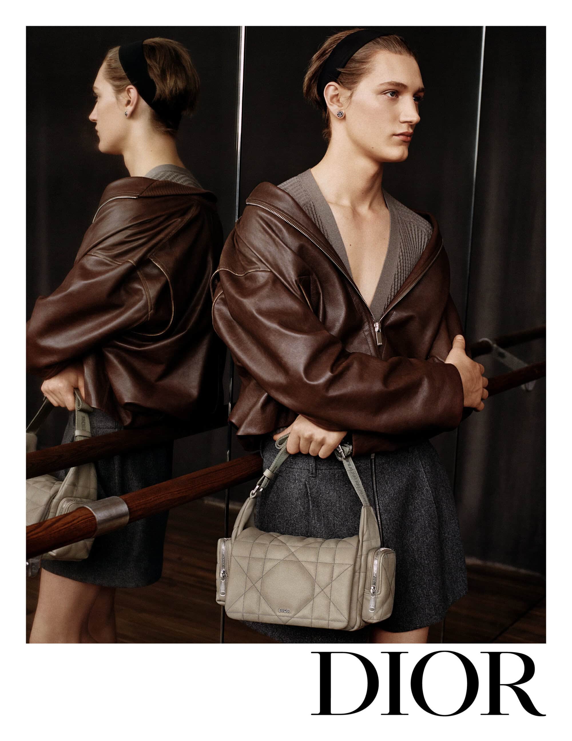 Fashion shot of a male model in a brown leather jacket holding a grey handbag next to a mirror with the Dior logo at the bottom.