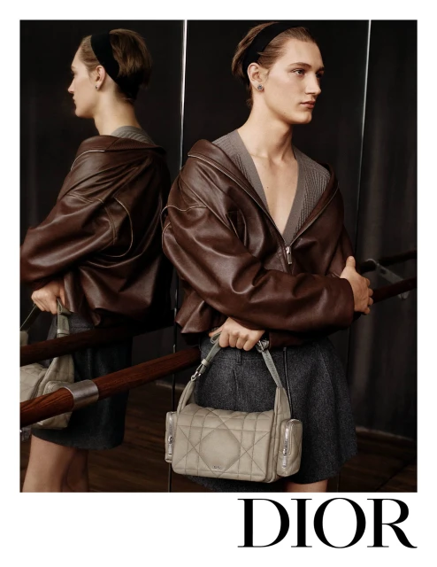 Fashion shot of a male model in a brown leather jacket holding a grey handbag next to a mirror with the Dior logo at the bottom.