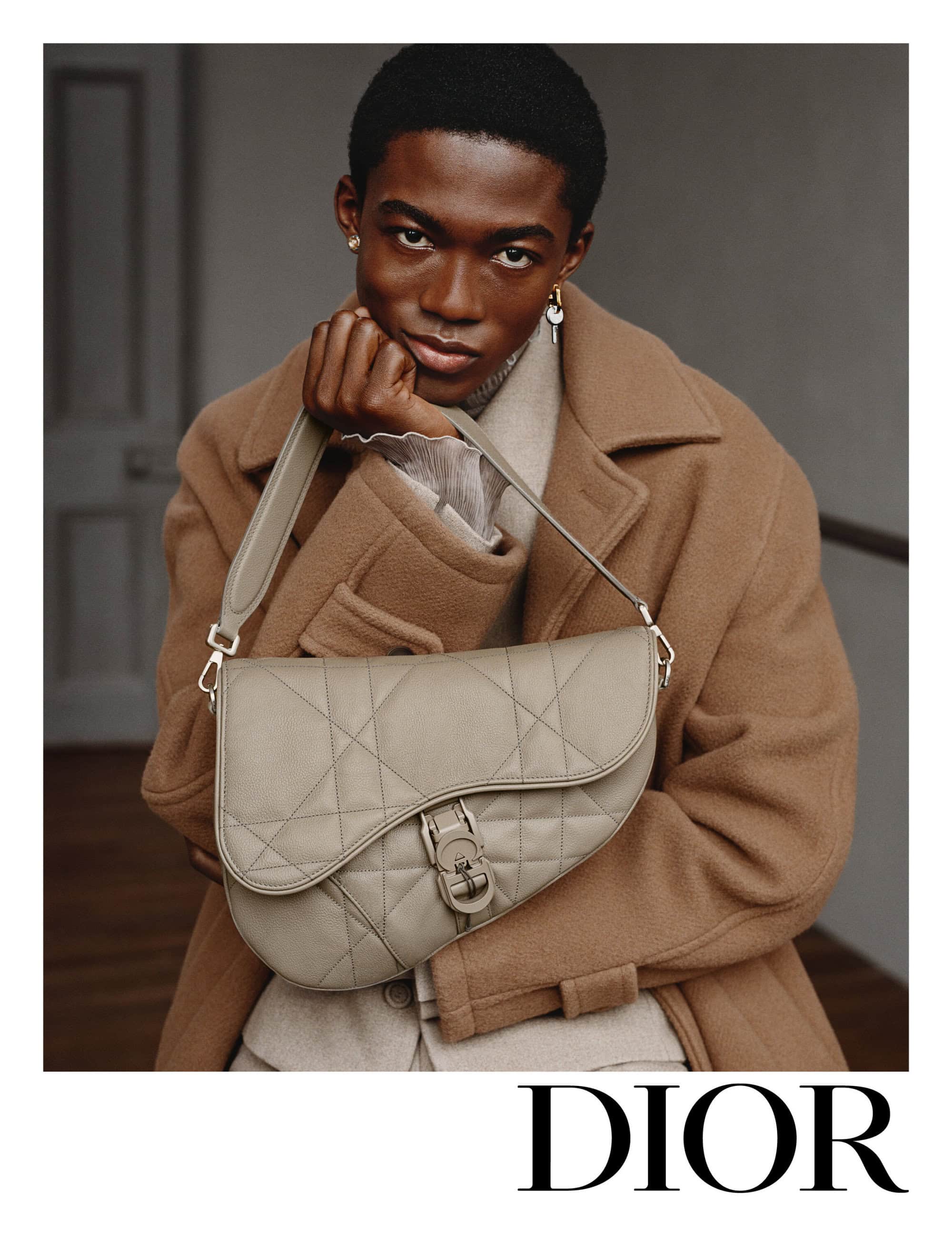 A man in a camel coat holding a quilted beige leather saddle bag in a studio setting