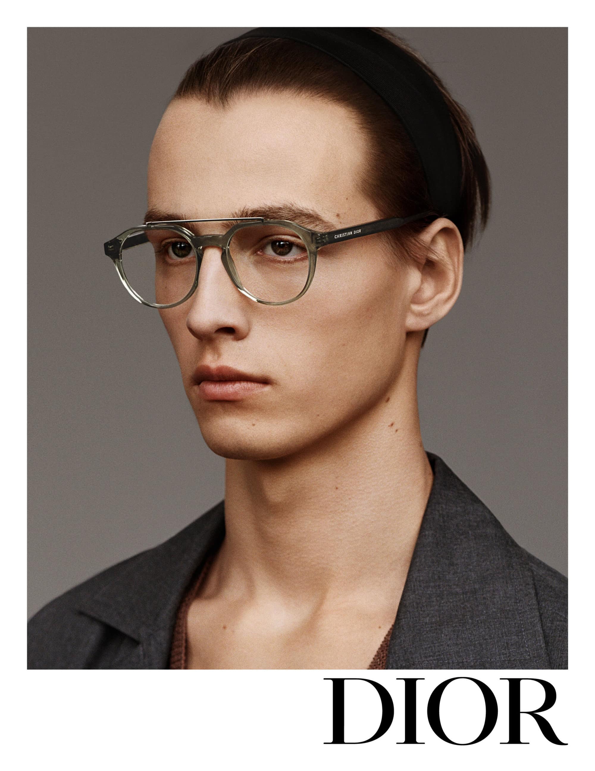 A light-skinned adult male model wears green-framed eyeglasses, a black headband, and a grey jacket against a neutral backdrop. Dior logo visible.