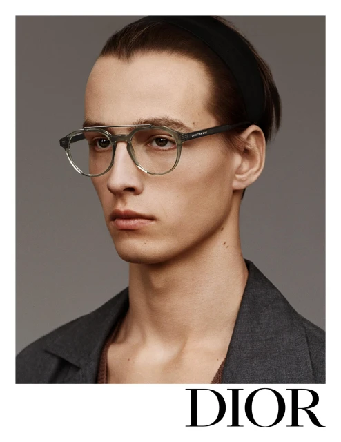 A light-skinned adult male model wears green-framed eyeglasses, a black headband, and a grey jacket against a neutral backdrop. Dior logo visible.