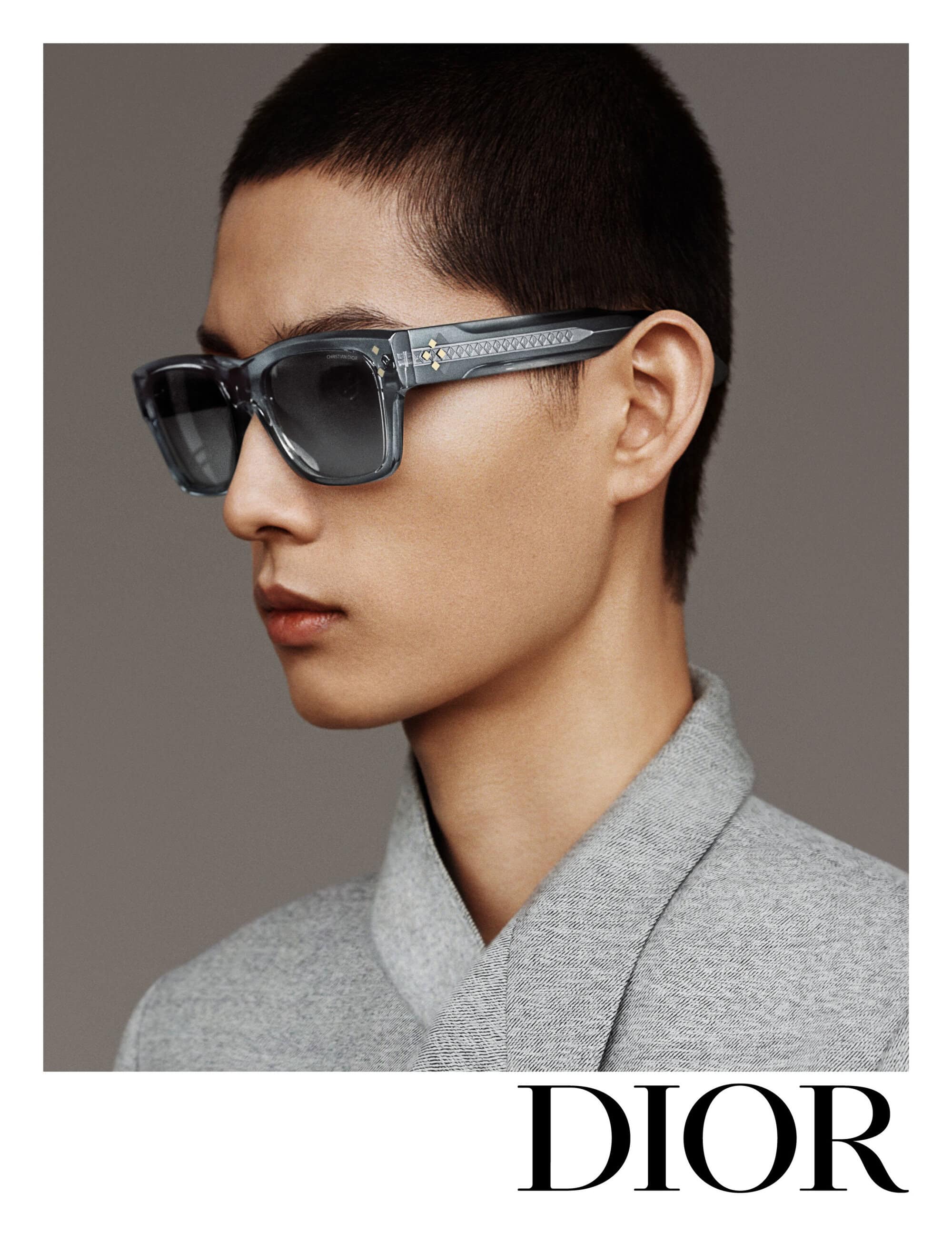 Close-up studio portrait of a young adult man wearing light blue designer sunglasses and a grey jacket against a neutral grey background.