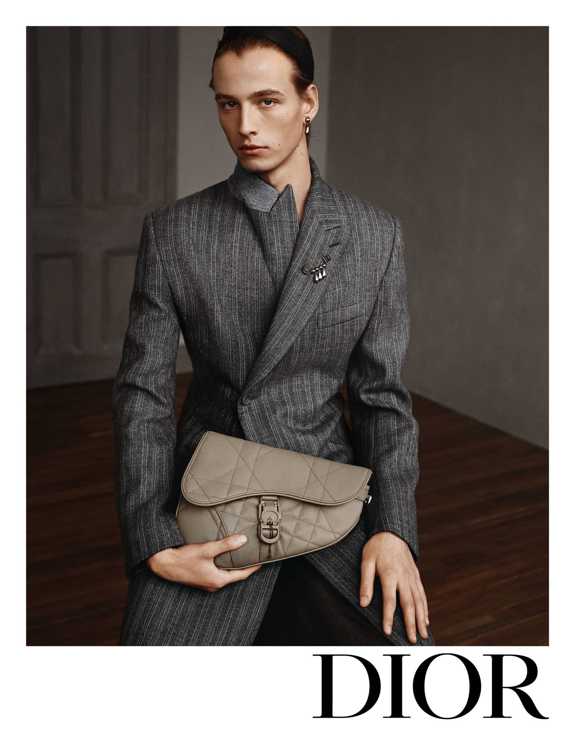 Man in grey suit holds saddle bag in indoor studio setting, Dior logo visible. Fashion editorial shot, indoor, neutral tones.