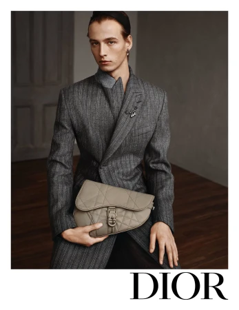 Man in grey suit holds saddle bag in indoor studio setting, Dior logo visible. Fashion editorial shot, indoor, neutral tones.