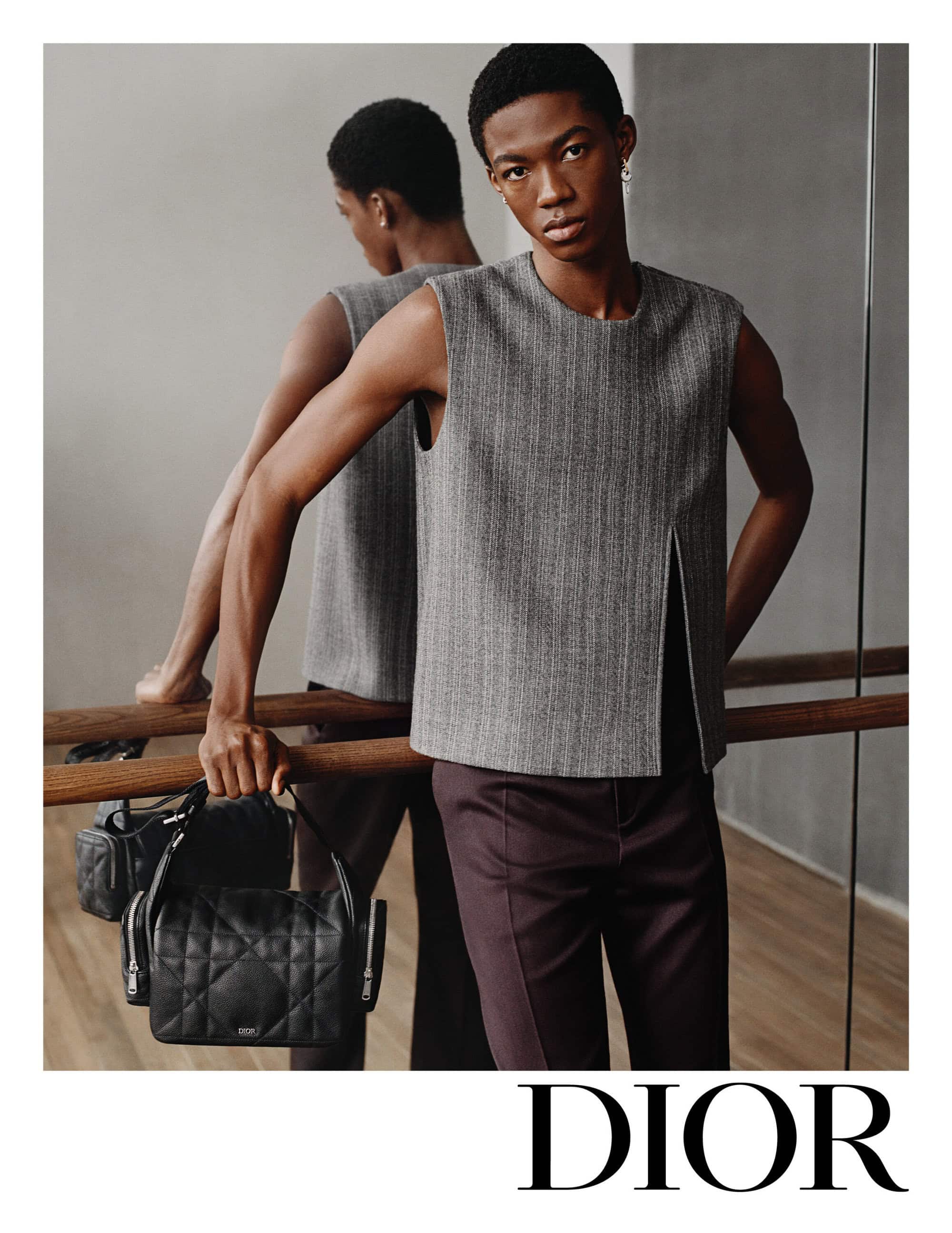 A young Black man wearing a grey sleeveless top and dark trousers holding a black quilted handbag against a mirror backdrop