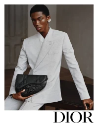 A young Black man wearing a light grey double-breasted suit holding a black leather saddle bag for Dior Men's Fall 2024 campaign