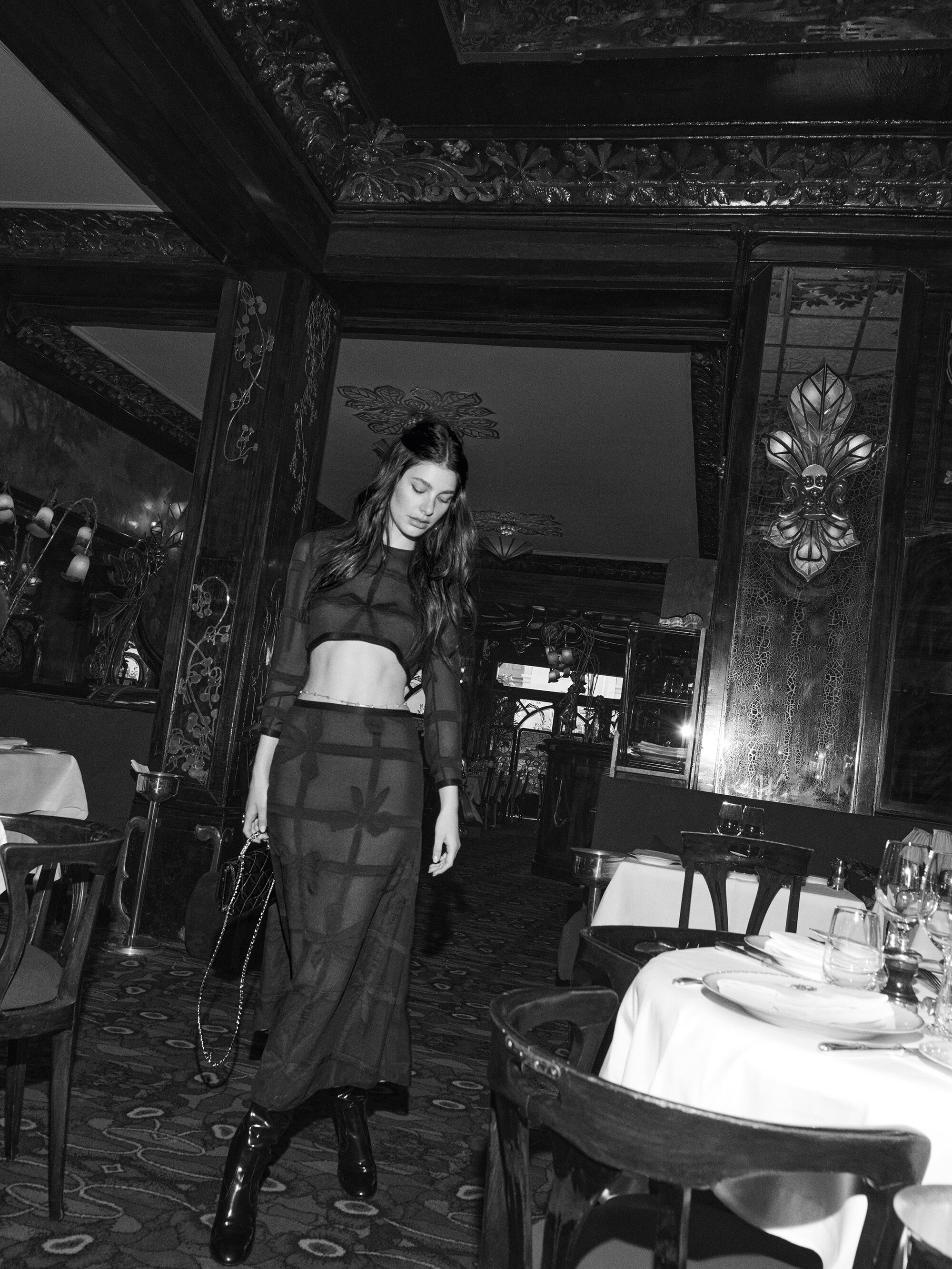 A woman in a sheer black outfit poses in a dimly lit restaurant with chairs, tables, and decor visible in the backdrop.