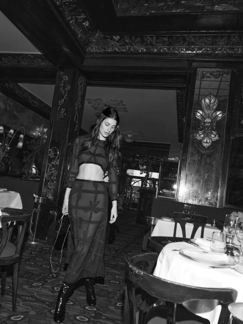 A woman in a sheer black outfit poses in a dimly lit restaurant with chairs, tables, and decor visible in the backdrop.