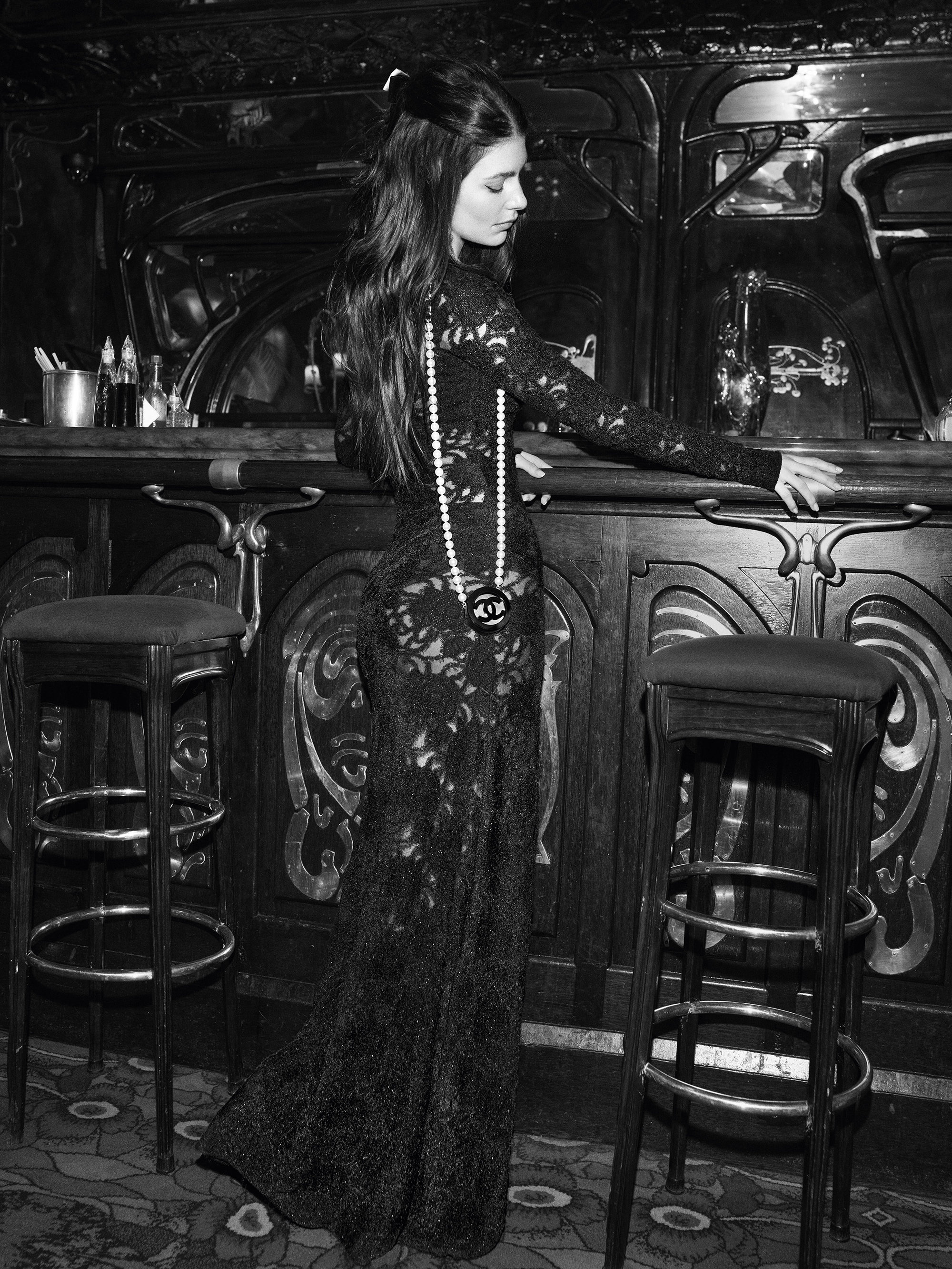 Black and white fashion photograph of a woman with long dark hair in an elegant black lace dress, standing near a dark ornate bar.