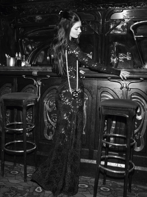 Black and white fashion photograph of a woman with long dark hair in an elegant black lace dress, standing near a dark ornate bar.