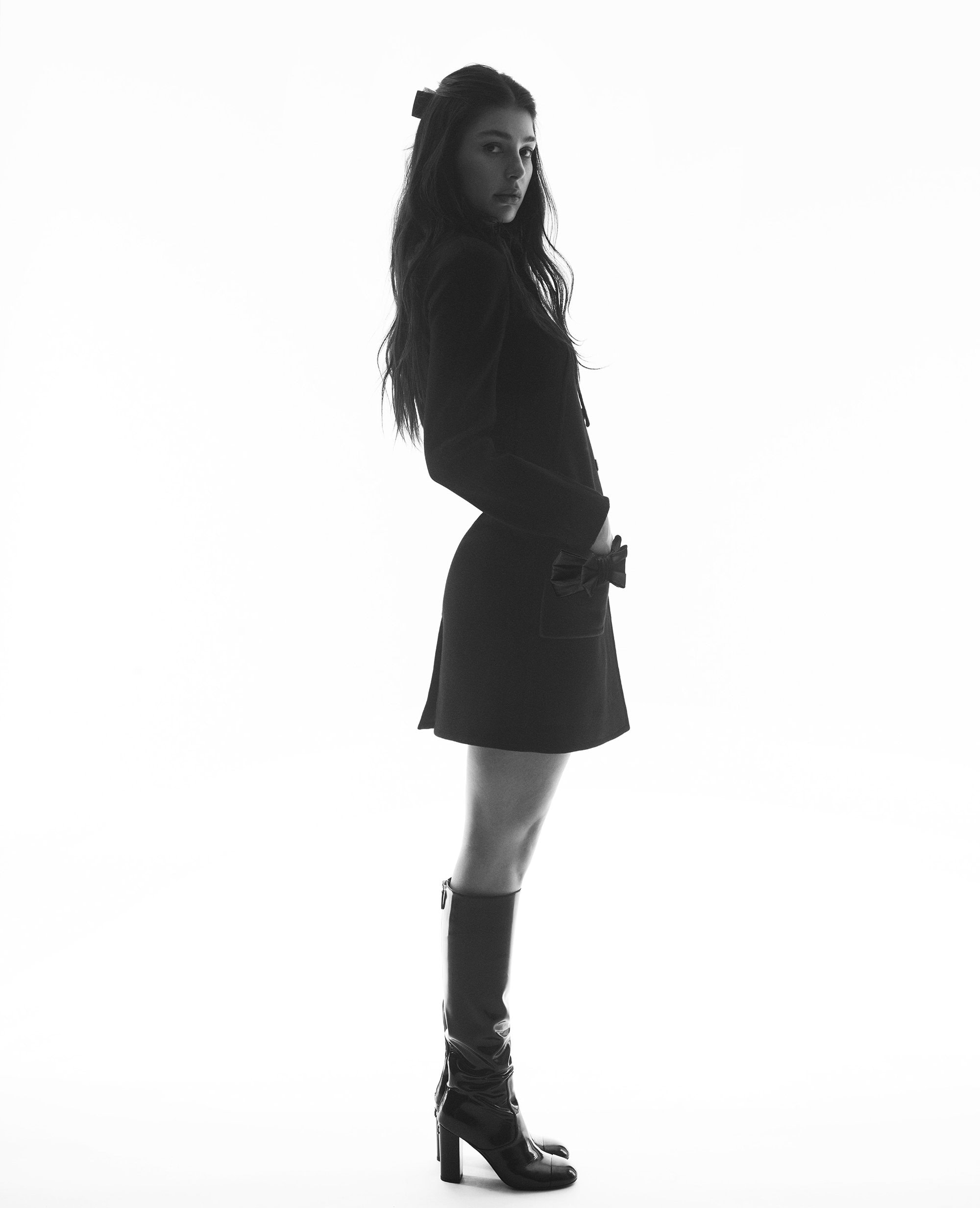 Model Camila Morrone wearing a dark coat and tall boots against a bright white background for Chanel Pre-Fall 2024