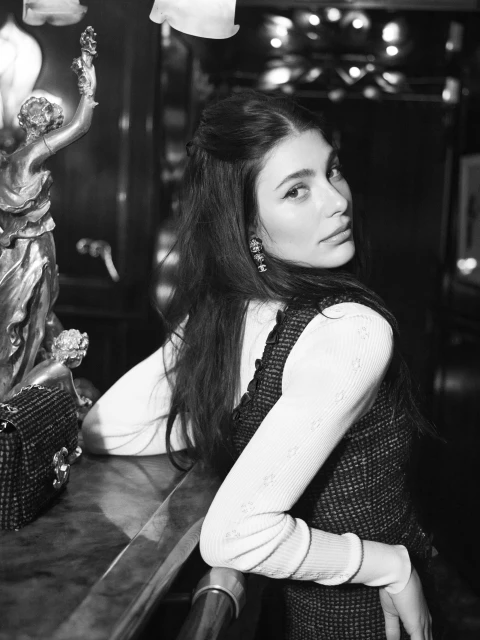 Monochrome portrait of a woman with dark hair, wearing a patterned top and white sleeves, leaning on a counter next to a small statue.