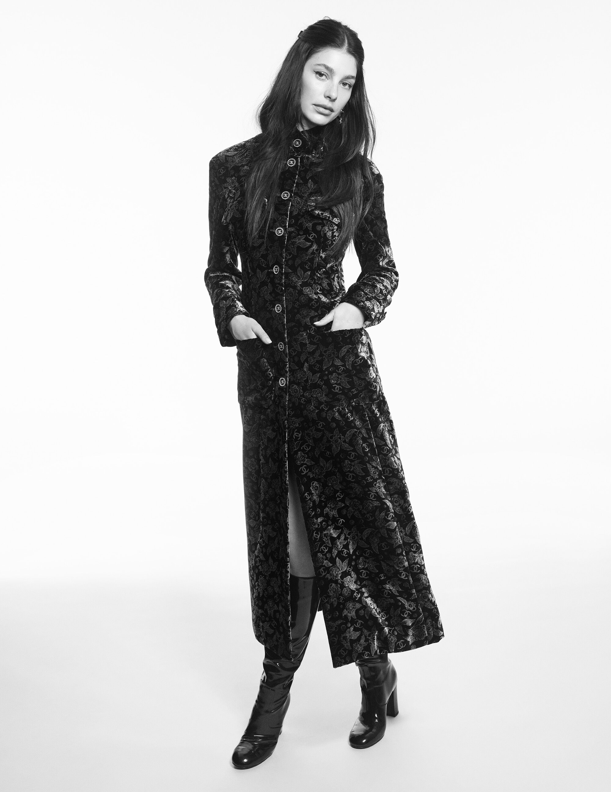 A woman in a long, patterned coat and shiny black boots stands against a bright background, posing for a fashion editorial shot.