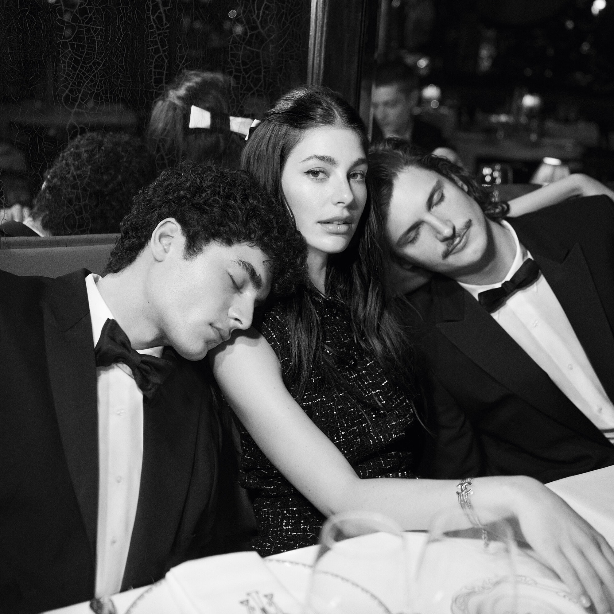 Black and white portrait of a woman flanked by two sleeping men wearing tuxedos, seated at a table in a restaurant setting.