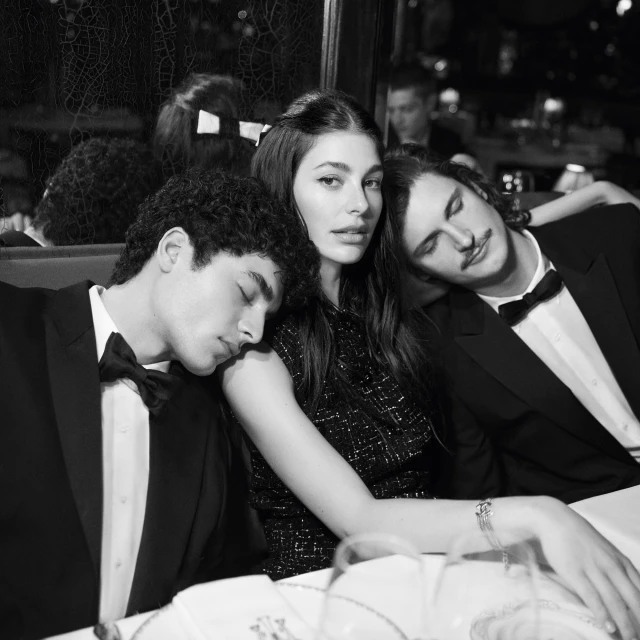 Black and white portrait of a woman flanked by two sleeping men wearing tuxedos, seated at a table in a restaurant setting.