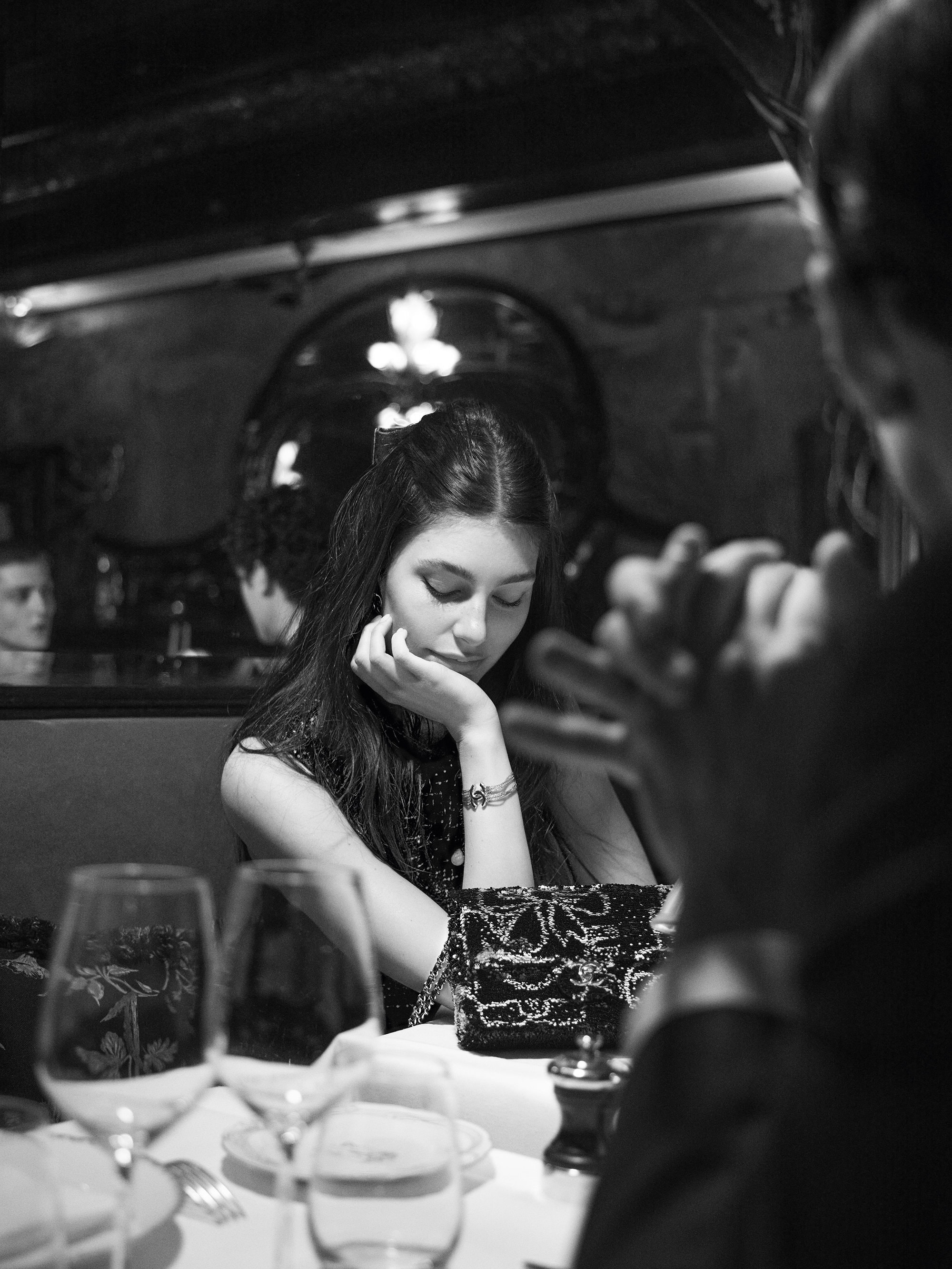 Black and white portrait of a woman seated at a table with wine glasses, a patterned purse, and another person's hands visible.