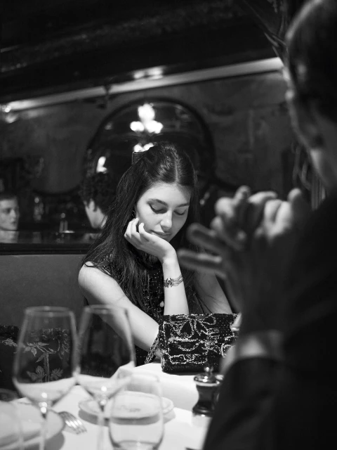 Black and white portrait of a woman seated at a table with wine glasses, a patterned purse, and another person's hands visible.