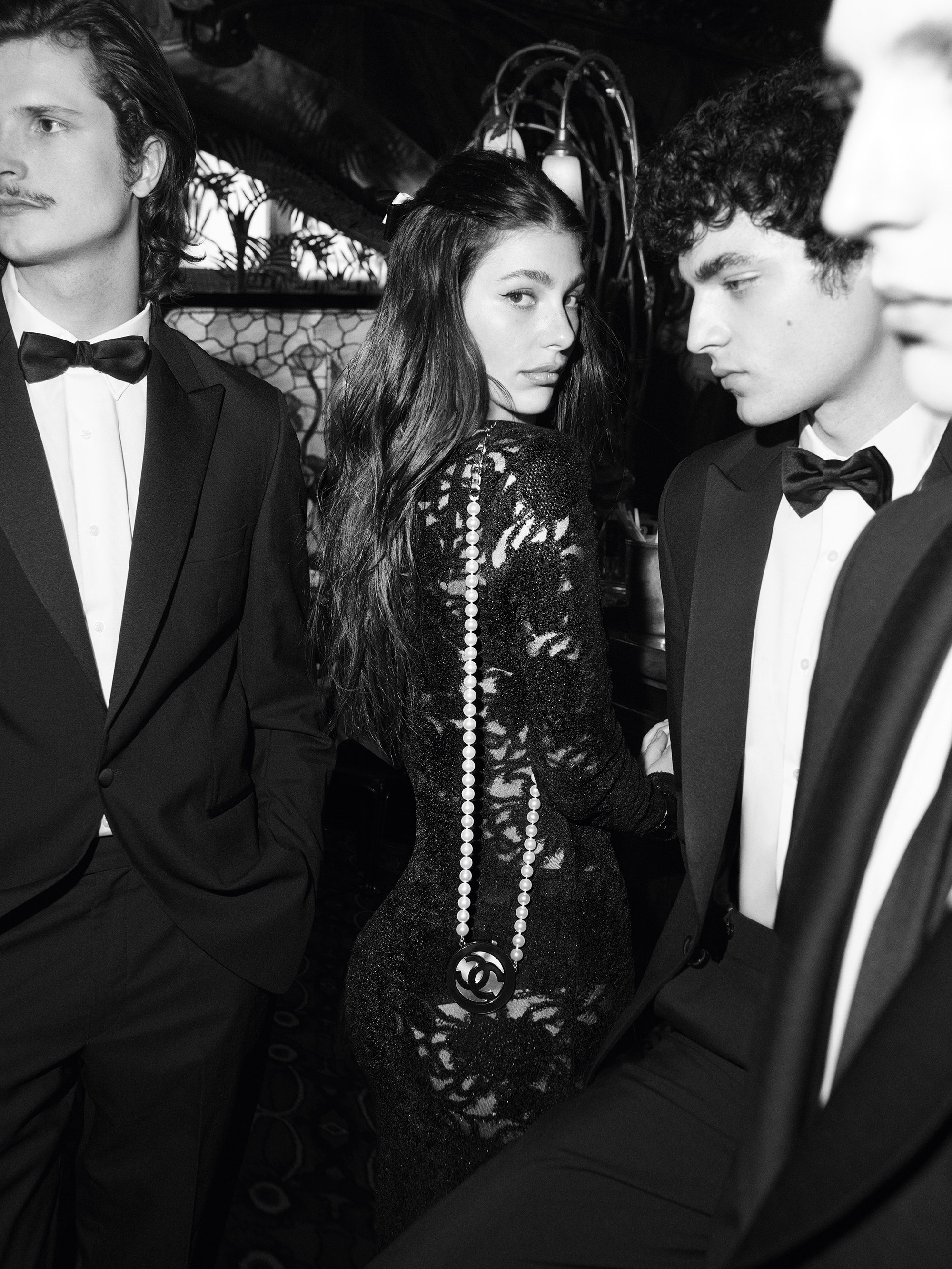 Black and white image of a woman in a black lace dress with pearl necklace and three men in black tuxedos at an event.