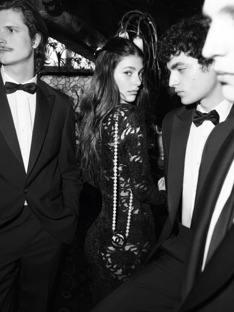 Black and white image of a woman in a black lace dress with pearl necklace and three men in black tuxedos at an event.