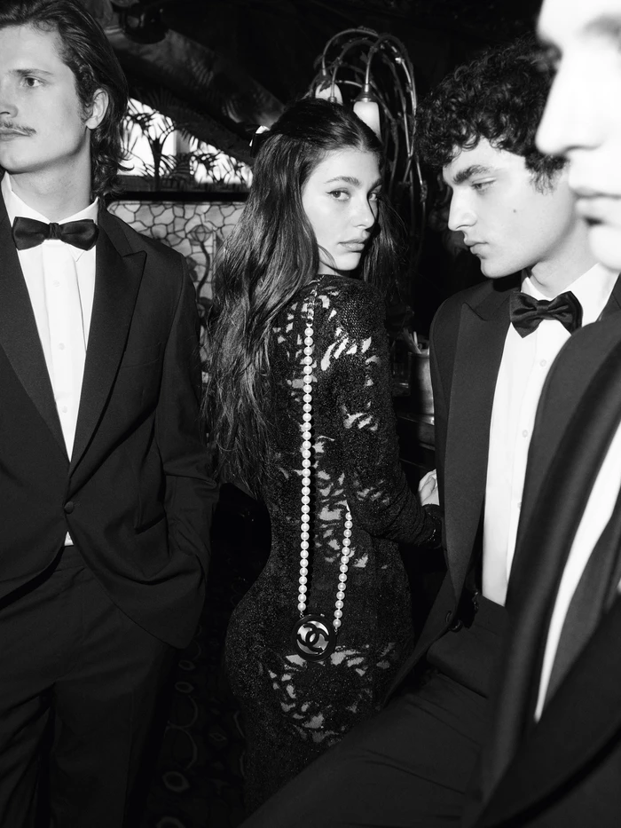 Camila Morrone in a black lace dress with a pearl-strapped bag, flanked by men in tuxedos in a black-and-white fashion campaign image.