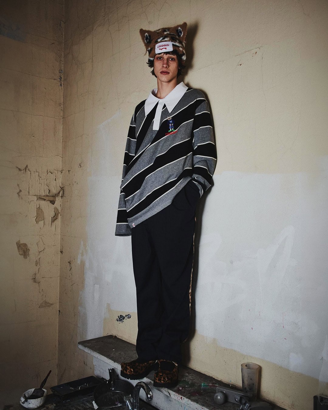 Stylish young adult poses indoors wearing a striped collared shirt, dark pants, and a quirky animal-eared hat with a distressed wall backdrop.