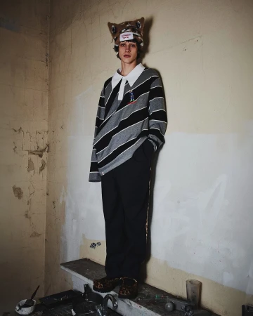Stylish young adult poses indoors wearing a striped collared shirt, dark pants, and a quirky animal-eared hat with a distressed wall backdrop.