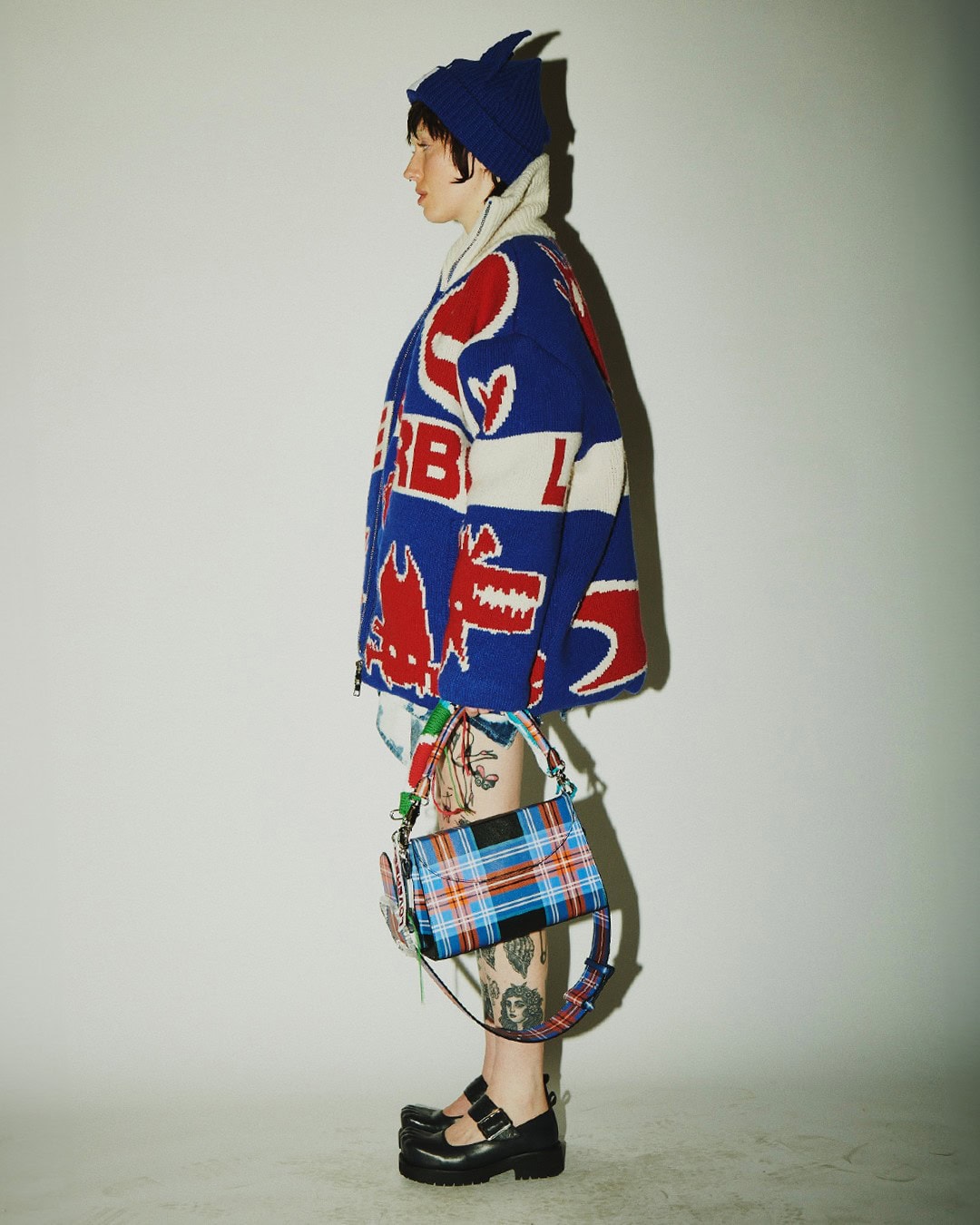 Stylish adult female in a blue and white knit outfit poses with a plaid purse indoors against a neutral background in a fashion shot.