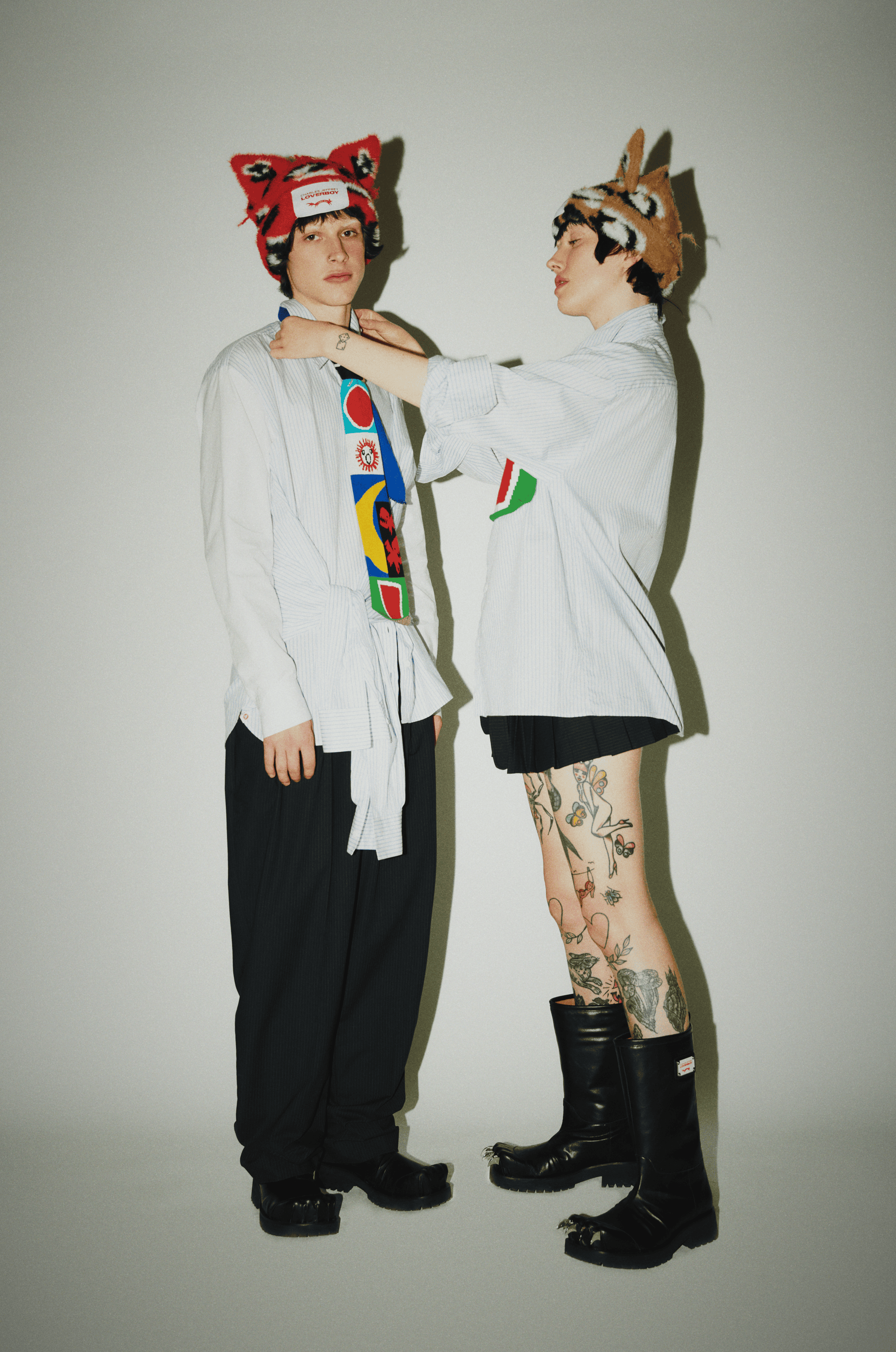Two people in a bright studio. One person adjusts a tie on the other. They wear matching quirky hats, light shirts, and dark pants.
