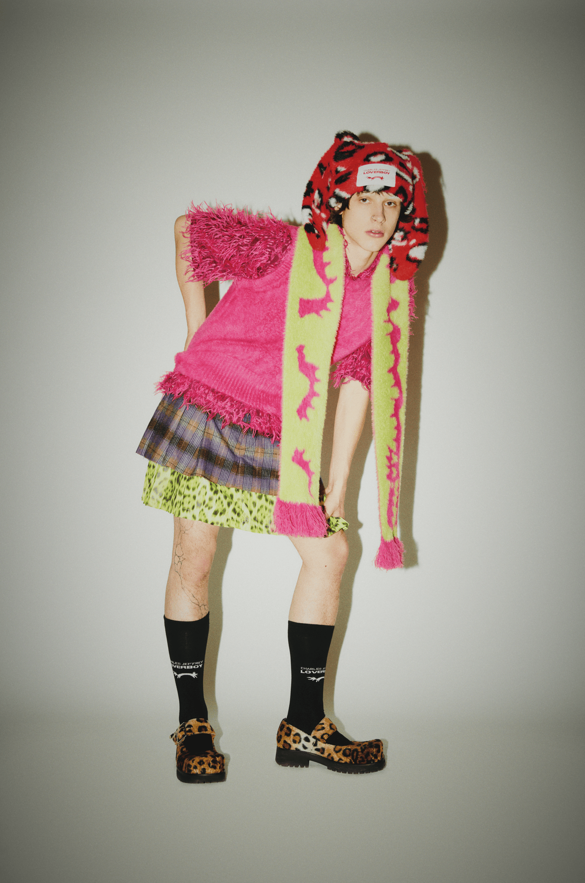 Person in a pink sweater and a plaid skirt with a patterned scarf and shoes posing in front of a plain white wall.