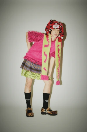 Person in a pink sweater and a plaid skirt with a patterned scarf and shoes posing in front of a plain white wall.