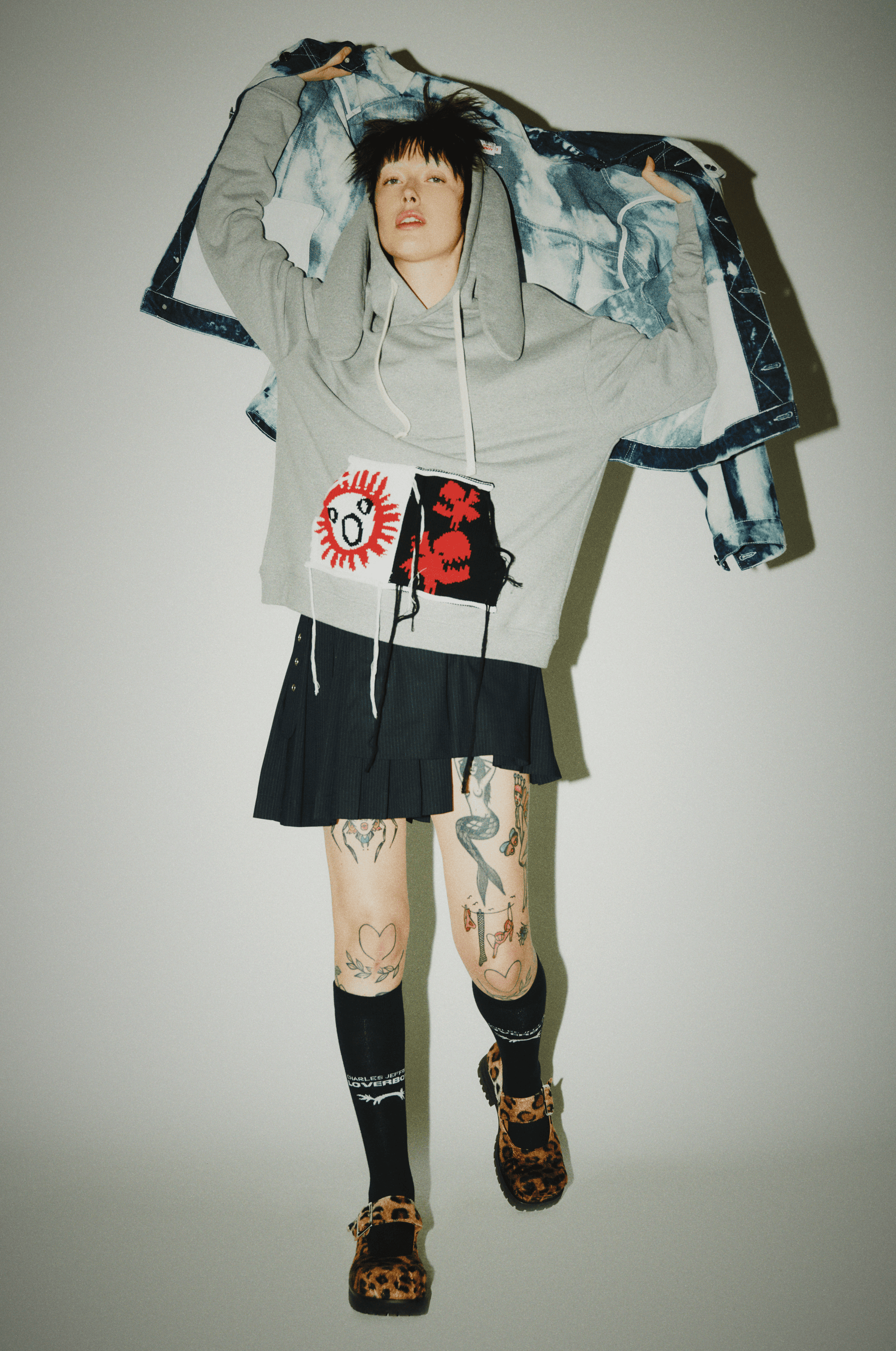Stylish model in a grey hoodie and black skirt poses in a studio, showcasing unique tattoos and leopard-print shoes against a plain background.
