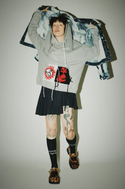 Stylish model in a grey hoodie and black skirt poses in a studio, showcasing unique tattoos and leopard-print shoes against a plain background.