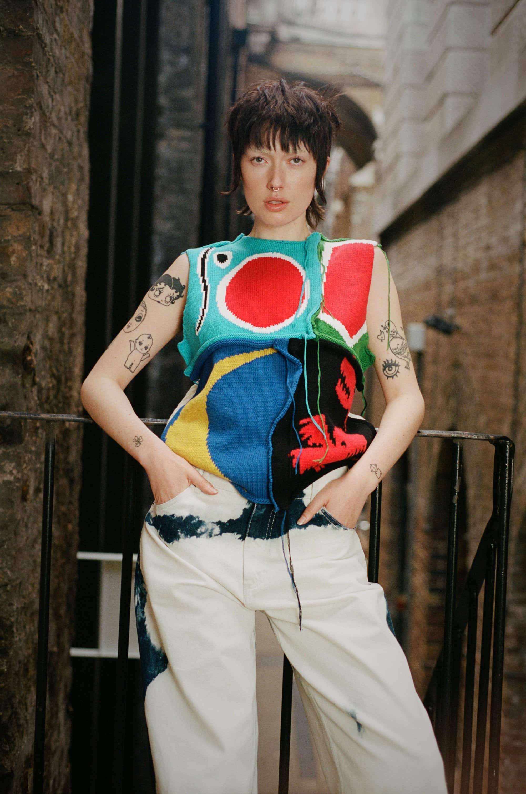 Woman in colorful knit top and white jeans poses in urban alleyway. Tattoos visible on arms. Eye-level shot with natural light.