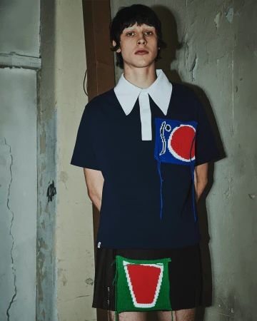 Young adult male fashion model poses in front of a weathered interior wall wearing a dark blue polo shirt and a black skirt.