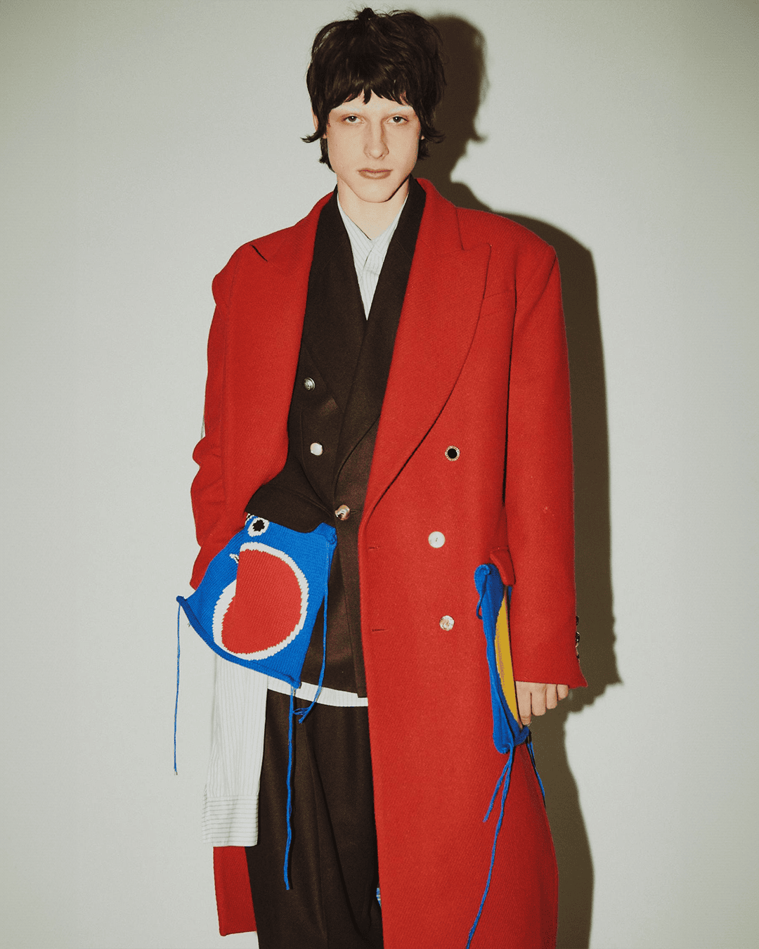 A person poses indoors in a red coat with attached colorful whimsical pouches, complemented by dark hair and a neutral background.