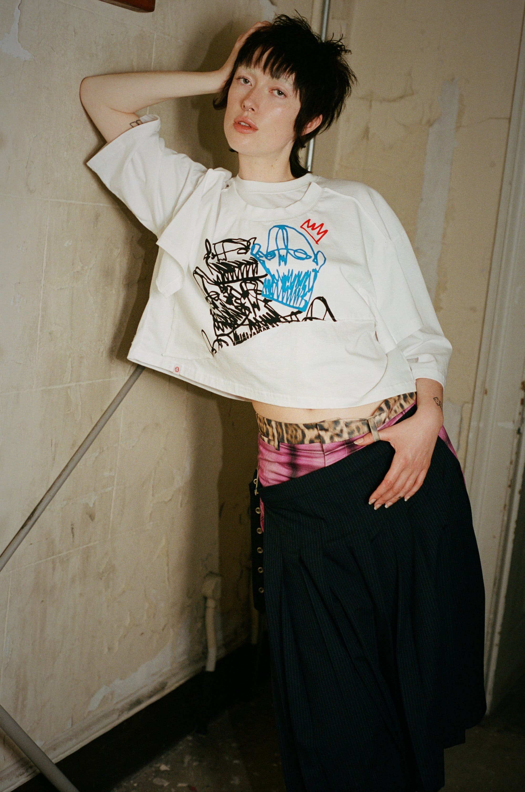 Woman in white graphic tee leans against a textured wall, wearing a skirt. Indoor studio shot with soft lighting.