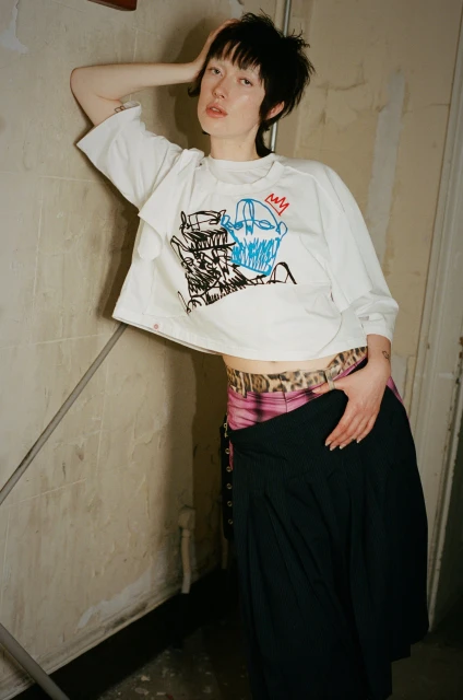 Woman in white graphic tee leans against a textured wall, wearing a skirt. Indoor studio shot with soft lighting.