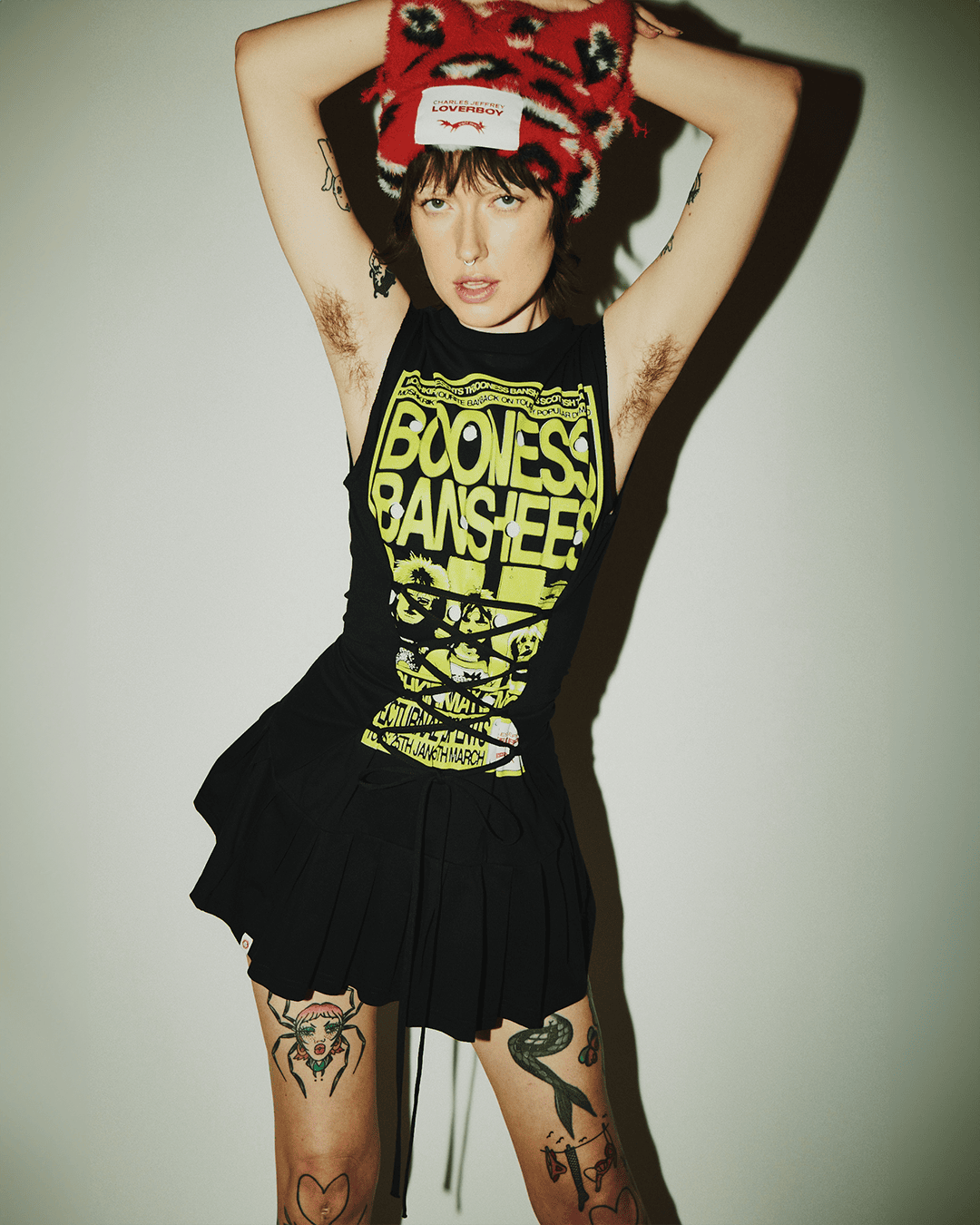 Portrait of a woman in a black dress with arm tattoos and armpit hair, wearing a patterned hat, and standing against a white wall.