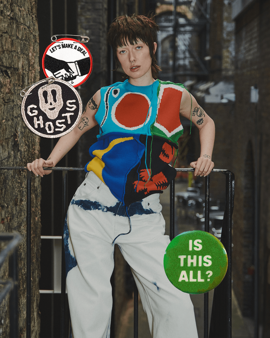 Portrait of a young adult female in a colorful knit vest and patterned pants, posing outdoors in a city alleyway with graphic pins.