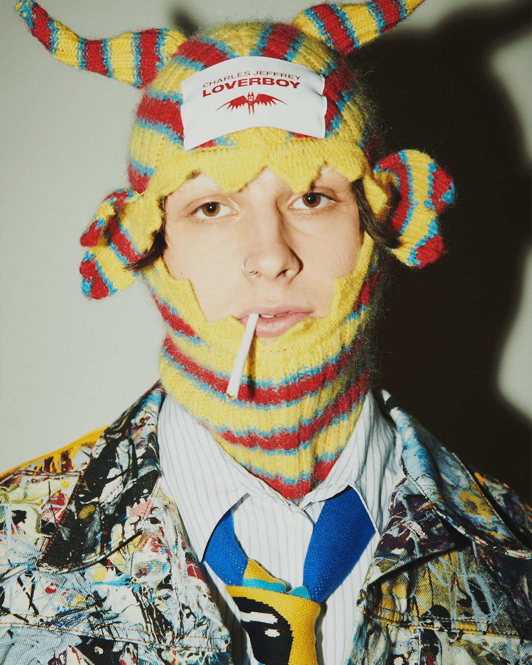 Portrait of a person wearing a yellow and red knitted balaclava-style hat with horns, blue tie, and a patterned jacket, holding a lollipop.