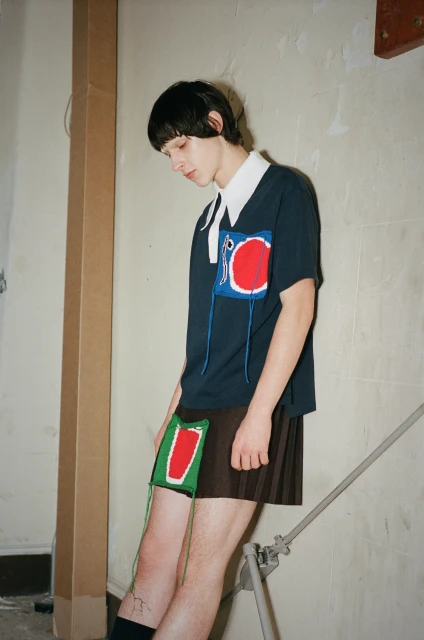 Young adult poses indoors wearing navy short-sleeve shirt, pleated skirt, and high socks for a fashion-forward studio portrait.