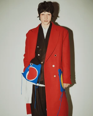 Model wearing a structured red coat over dark layers with distinctive blue bag accessories for Charles Jeffrey Loverboy Fall 2024