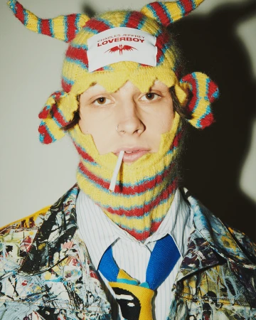 Model wearing a yellow and red striped horned beanie hat with a white branded label and a paint-splattered jacket.