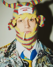 Model wearing a yellow and red striped horned beanie hat with a white branded label and a paint-splattered jacket.