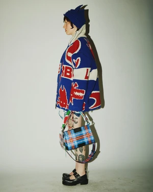 Model in profile wearing a blue patterned knit jacket and matching hat, carrying a plaid handbag against a neutral background.