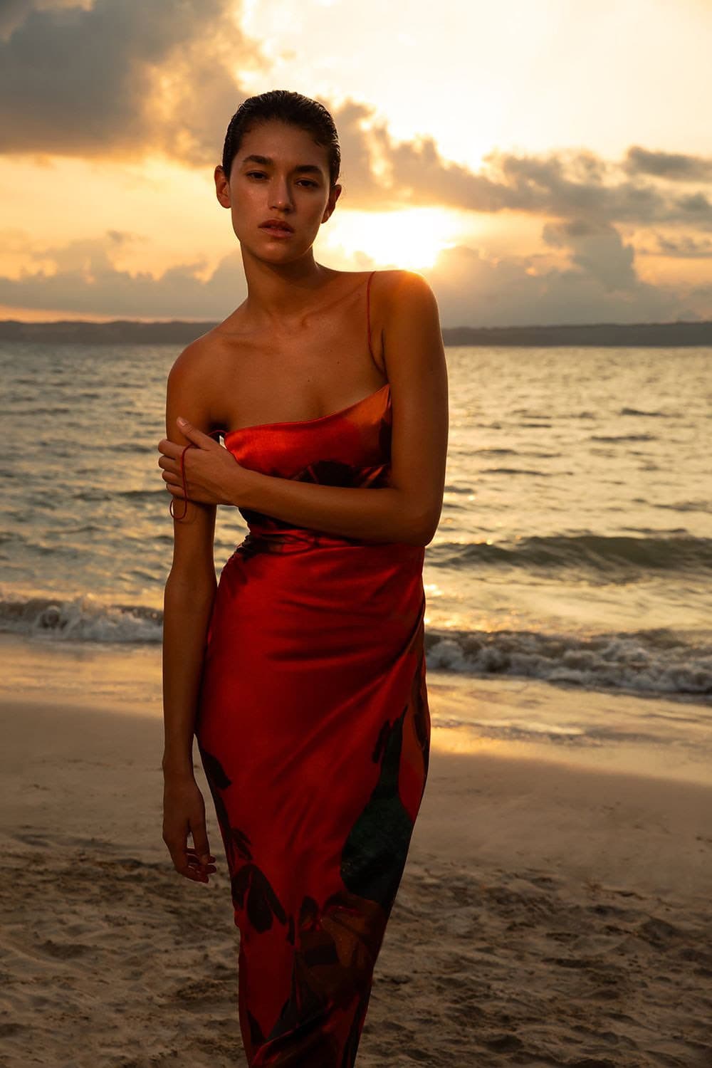 Model in red silk dress standing on sandy beach at sunset for SIR. Pre-Fall 2024 campaign
